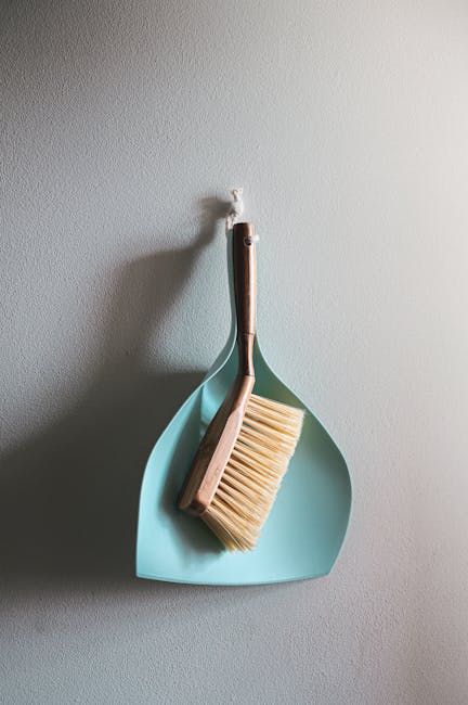 A light blue dustpan hanging on a white wall with a wooden-handled broom and a yellow-bristled brush placed inside it. The cleaning tools are neatly arranged and appear clean and well-maintained, suit