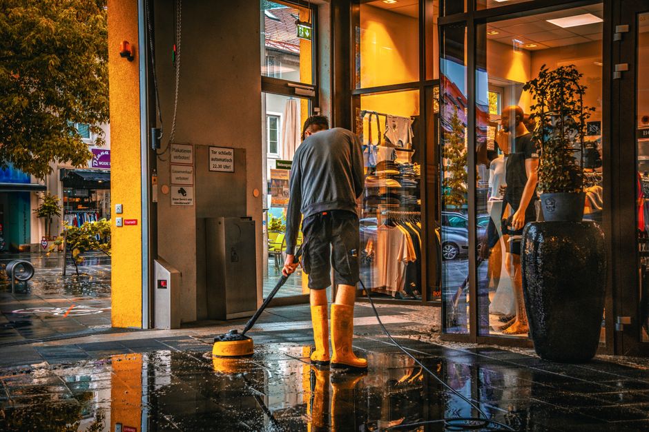 A person wearing yellow rubber boots and casual clothing is engaged in surface cleaning outside a commercial retail store, using a power scrubber on the wet pavement. The store’s large glass windows