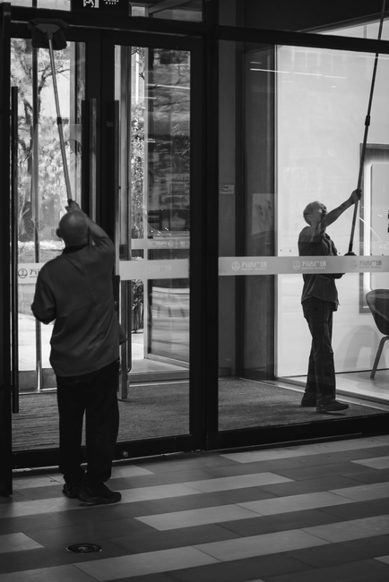 A person wearing yellow rubber boots and casual clothing is engaged in surface cleaning outside a commercial retail store, using a power scrubber on the wet pavement. The store’s large glass windows